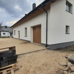 Nowoczesny dom jednorodzinny z jasną elewacją, brązową bramą garażową i ułożoną kostką brukową na podjeździe, widok z poziomu gruntu z widocznymi paletami z kostką i kamieniami na pierwszym planie.