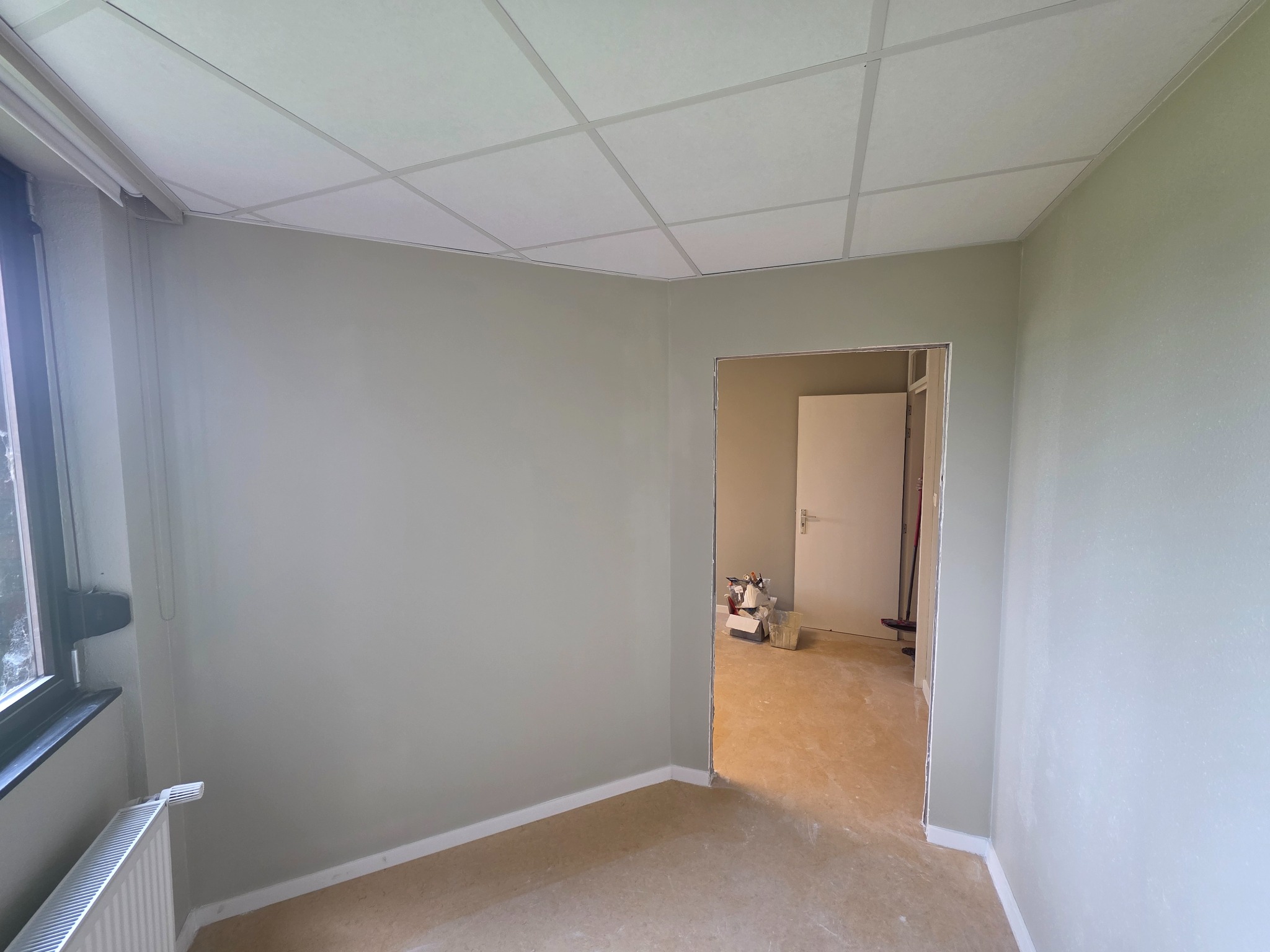 Wnętrze pomalowanego na szaro pokoju z kasetonowym sufitem, widok na sąsiednie pomieszczenie z drzwiami i narzędziami malarskimi. Widoczny grzejnik.