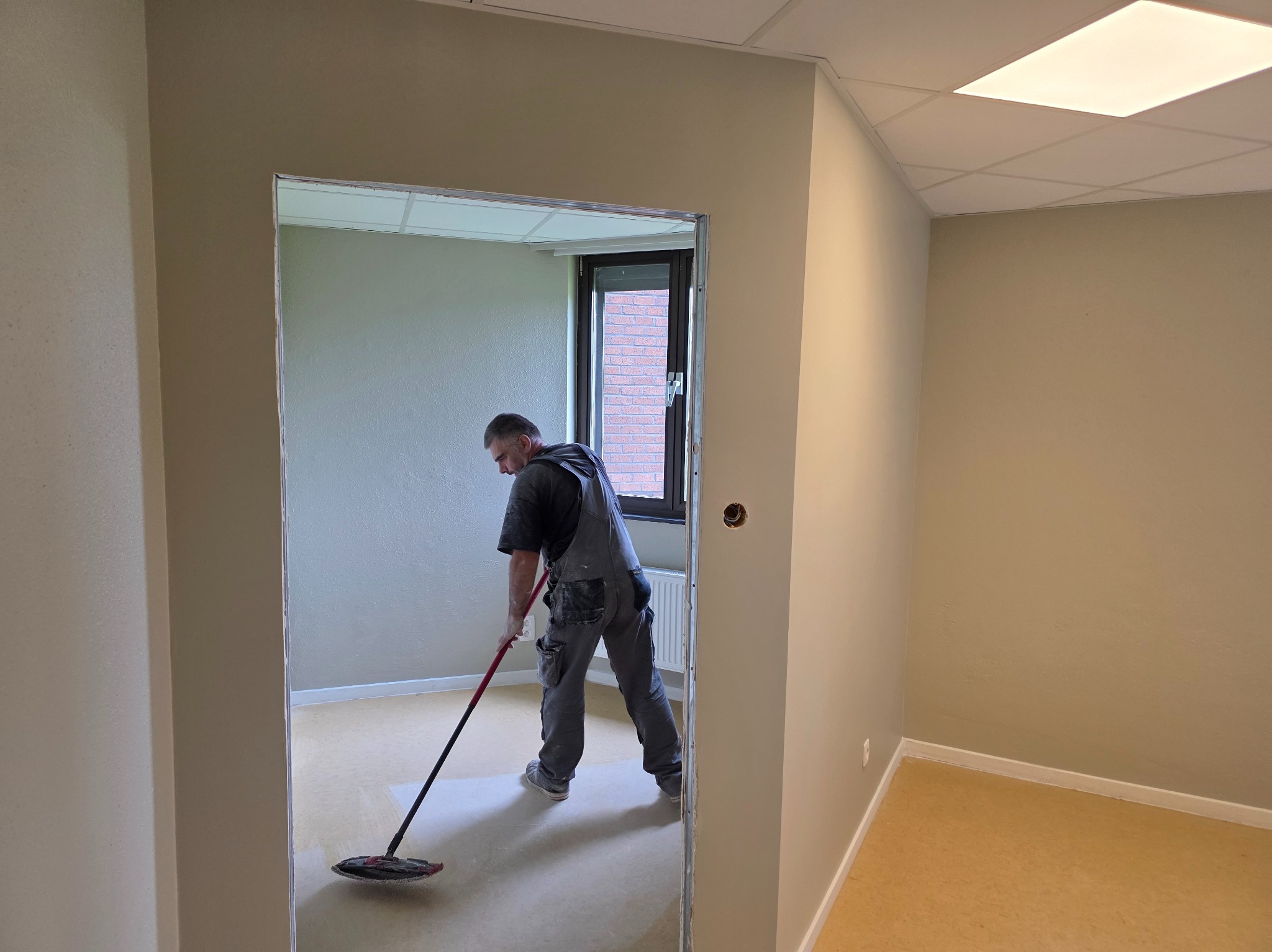 Mężczyzna w kombinezonie maluje podłogę w pokoju biurowym za pomocą mopa. Widoczne okno i grzejnik. Praca w trakcie wykonywania, jasne oświetlenie.