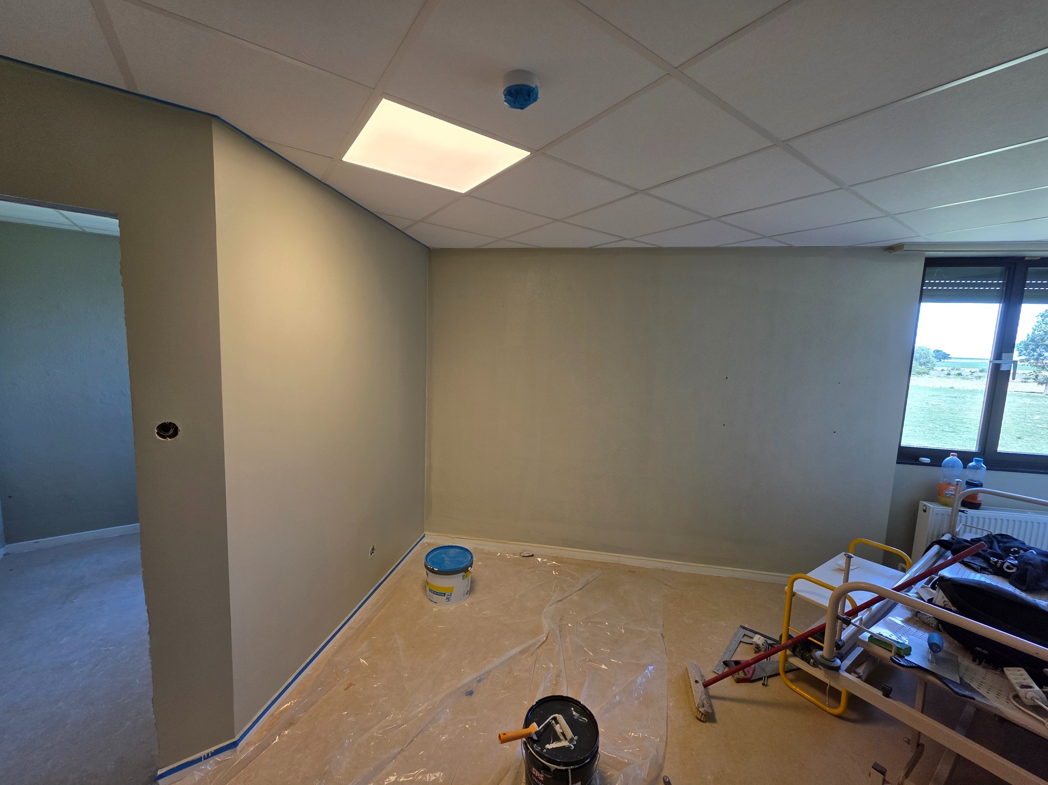 Wnętrze po malowaniu ścian na szaro, z widocznymi narzędziami malarskimi, wiaderkami farby i zabezpieczoną podłogą folią. Okno z widokiem na zewnątrz.