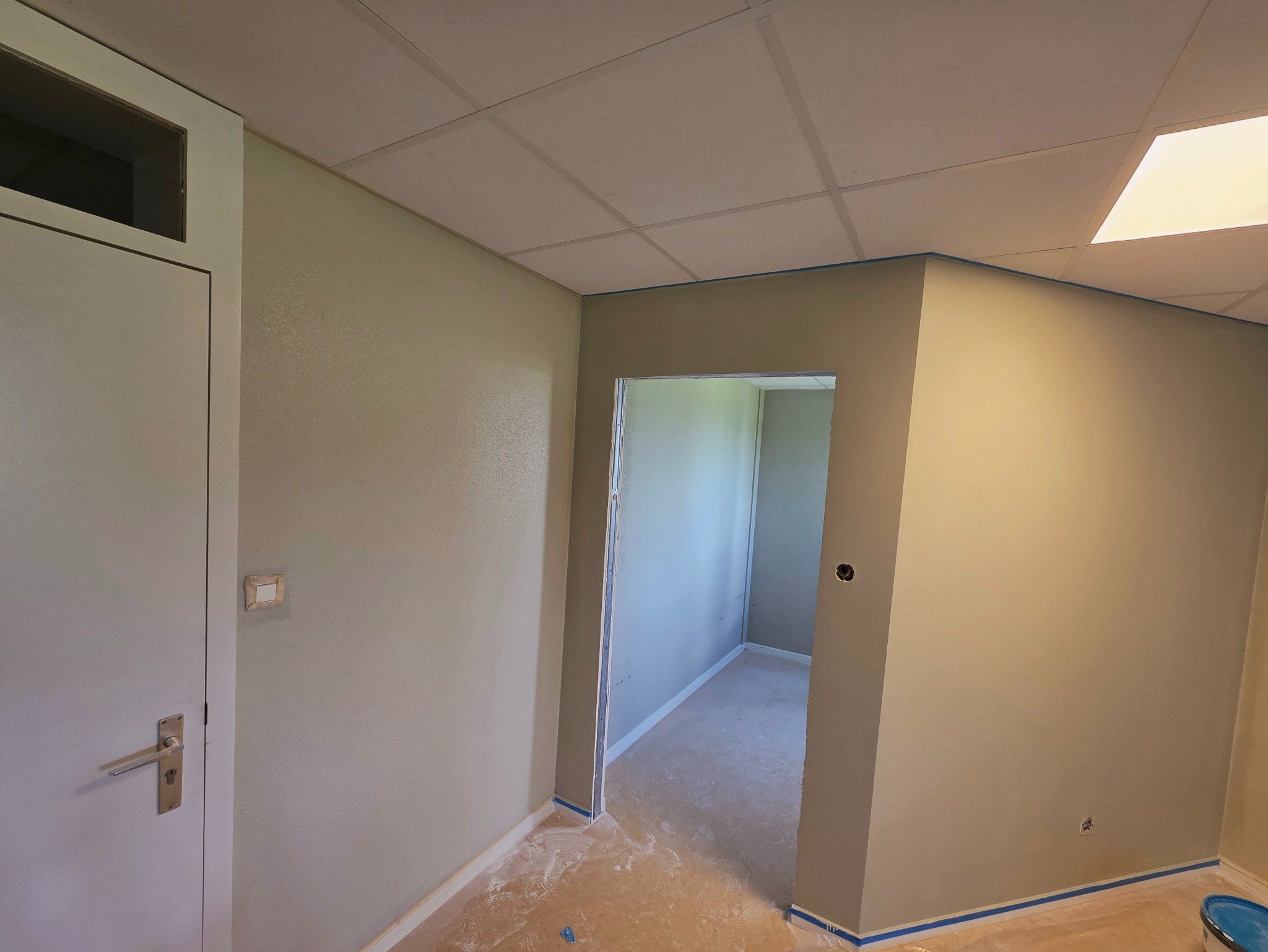 Świeżo pomalowane wnętrze biura w odcieniach szarości, z zabezpieczonymi listwami przypodłogowymi niebieską taśmą malarską i widocznym fragmentem sufitu kasetonowego.