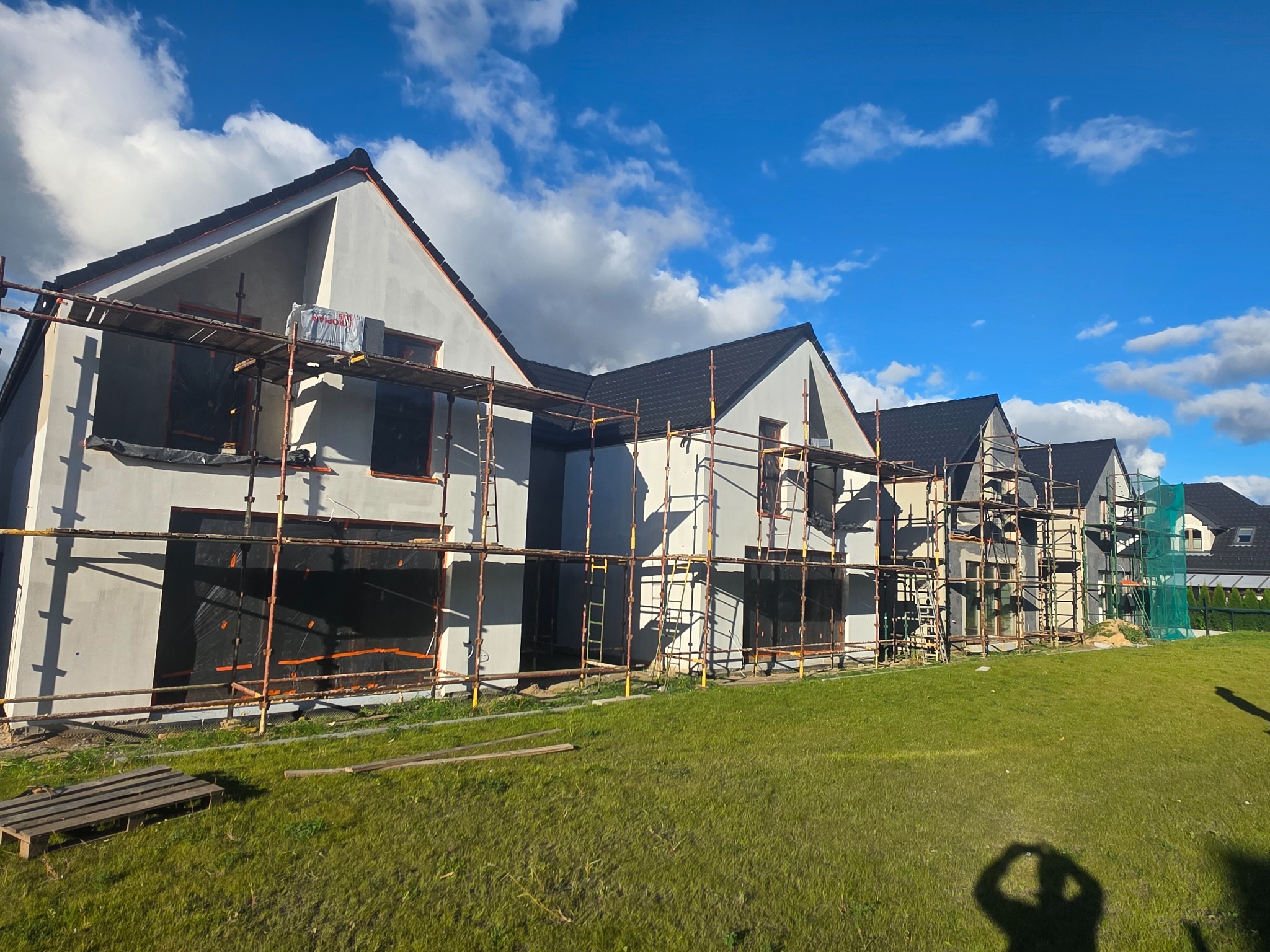 Nowoczesne domy w zabudowie szeregowej w trakcie prac elewacyjnych. Widoczne rusztowania i częściowo otynkowane ściany. Błękitne niebo z chmurami w tle.