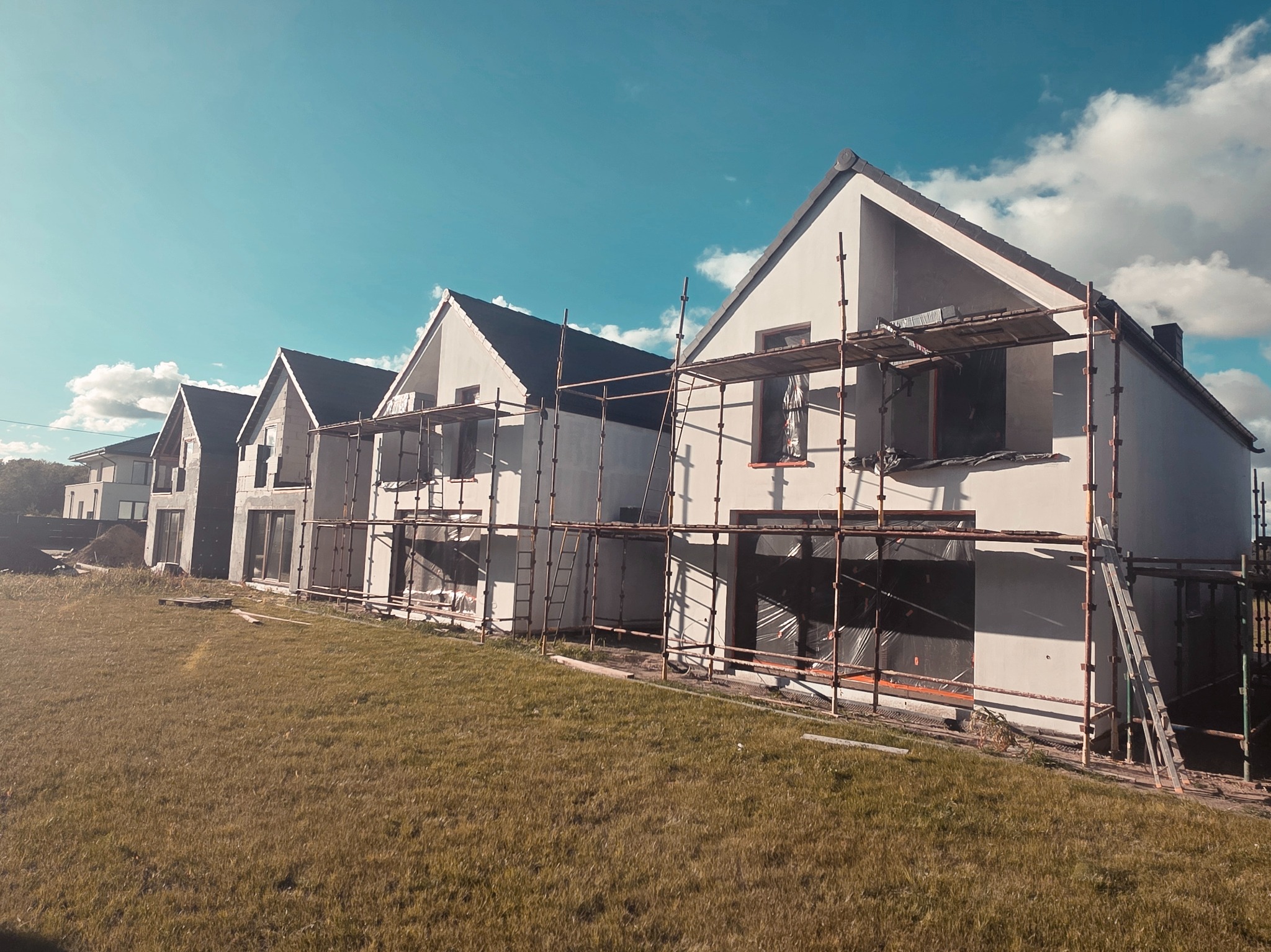 Rząd domów jednorodzinnych w trakcie tynkowania elewacji, z widocznymi rusztowaniami i przygotowanym terenem wokół. Słoneczny dzień, błękitne niebo.