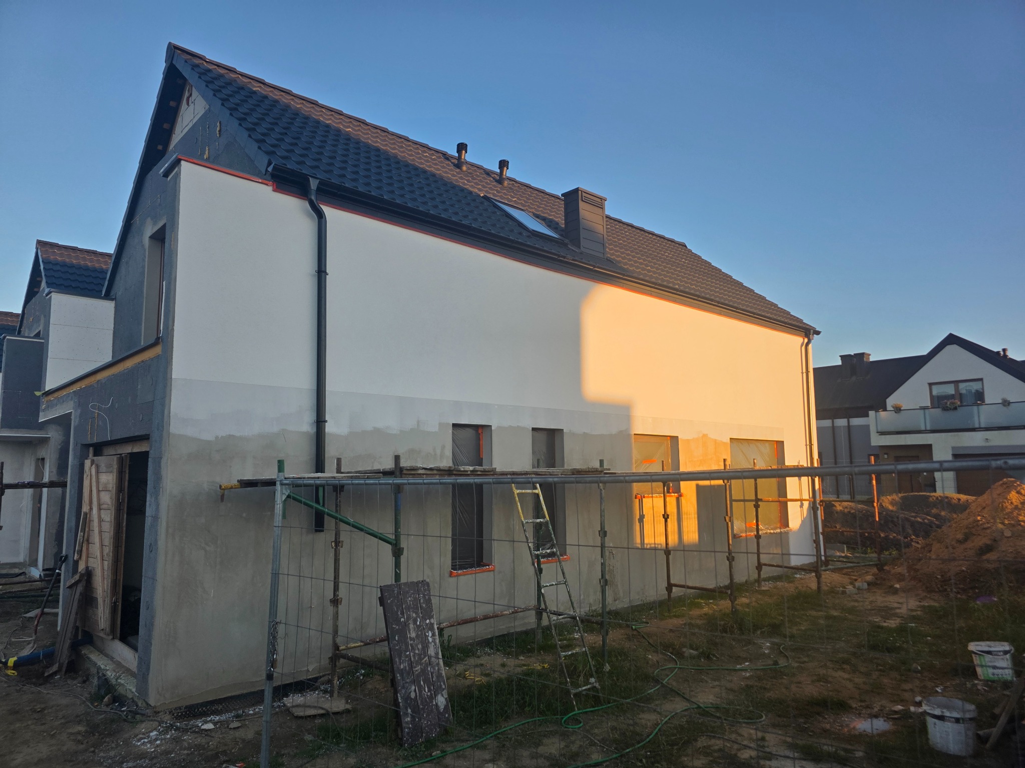 Dom w trakcie prac elewacyjnych: ocieplanie i tynkowanie. Widoczne rusztowania, fragmenty otynkowanej ściany i niezabezpieczone okna. Częściowo zachmurzone niebo.