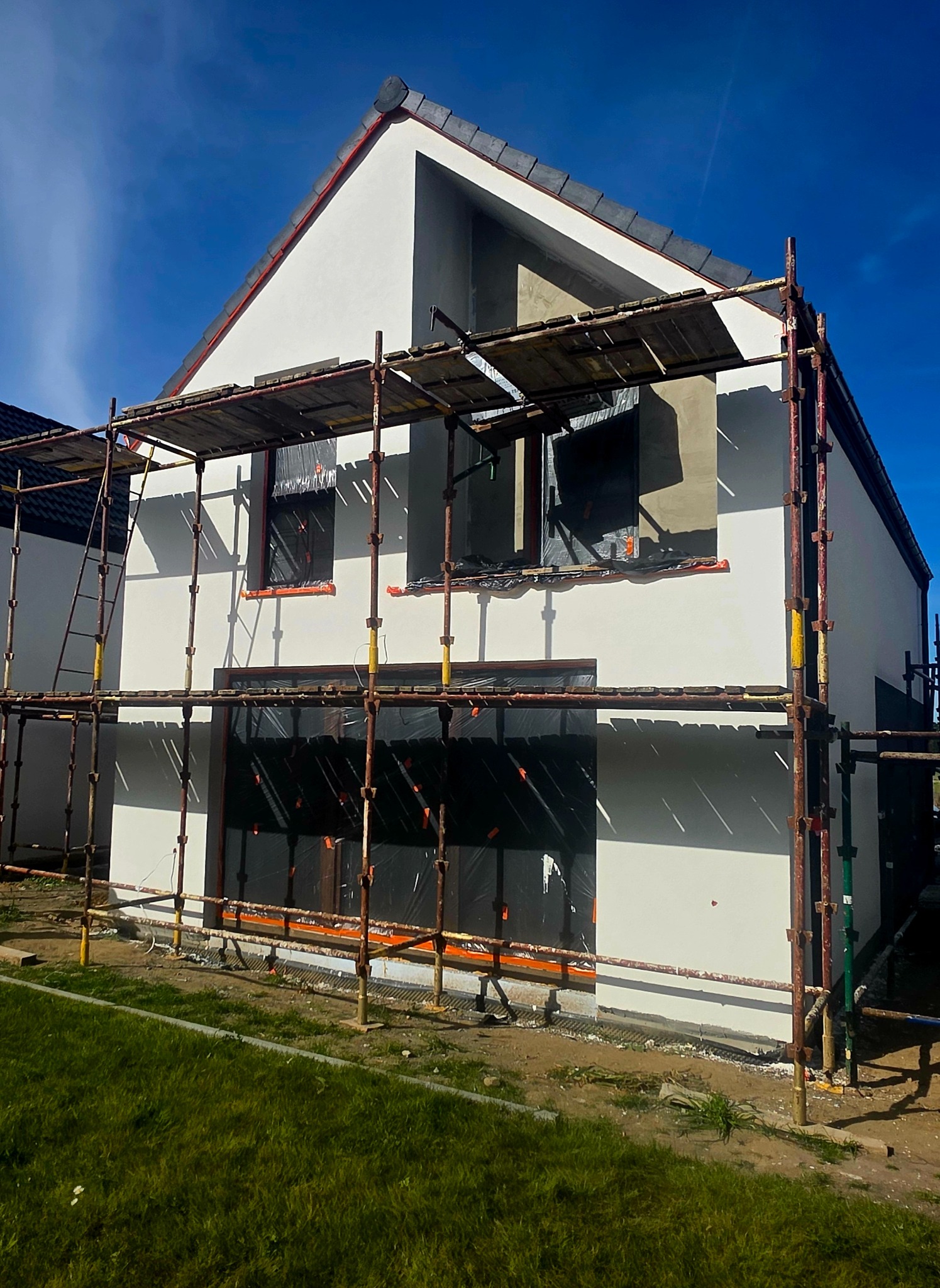 Biała elewacja domu w trakcie prac wykończeniowych. Widoczne rusztowania, zabezpieczone okna i fragmenty surowego tynku. Dom z ciemną dachówką na tle błękitnego nieba.