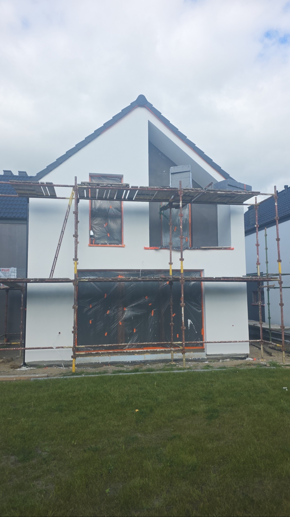 Dom w trakcie prac elewacyjnych: rusztowania, okna zabezpieczone folią, fragment otynkowanej ściany. Widoczne fragmenty sąsiednich budynków i trawnik.