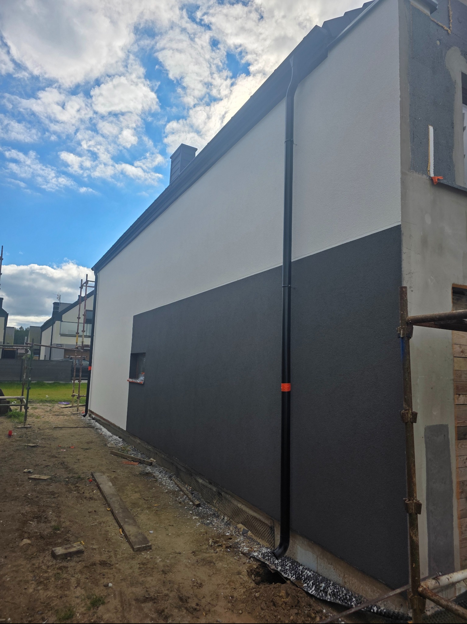 Dwukolorowa elewacja domu w trakcie wykończenia, z widocznymi rusztowaniami i fragmentami ocieplenia. Część ściany pomalowana na szaro, część na biało.