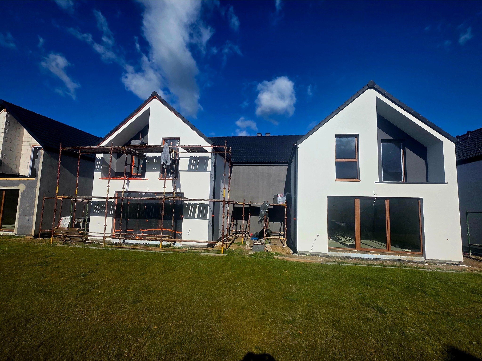 Dwukondygnacyjny dom w trakcie prac elewacyjnych, z rusztowaniami i fragmentami ocieplenia, na tle błękitnego nieba z chmurami. Widok z trawnika.