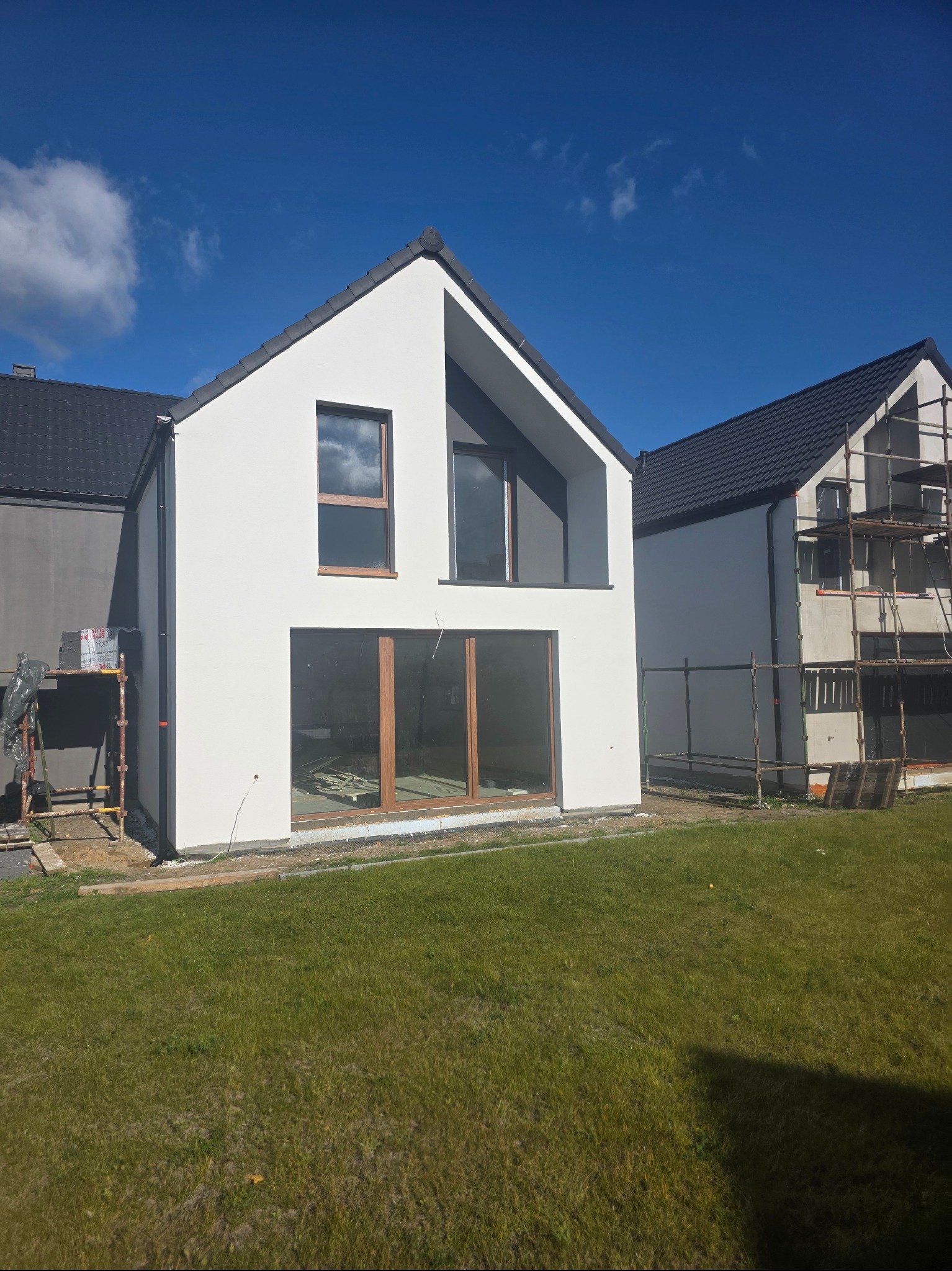 Biała elewacja domu jednorodzinnego z drewnianą stolarką okienną, widok z zewnątrz na tle błękitnego nieba. Na sąsiednim budynku widoczne rusztowania.