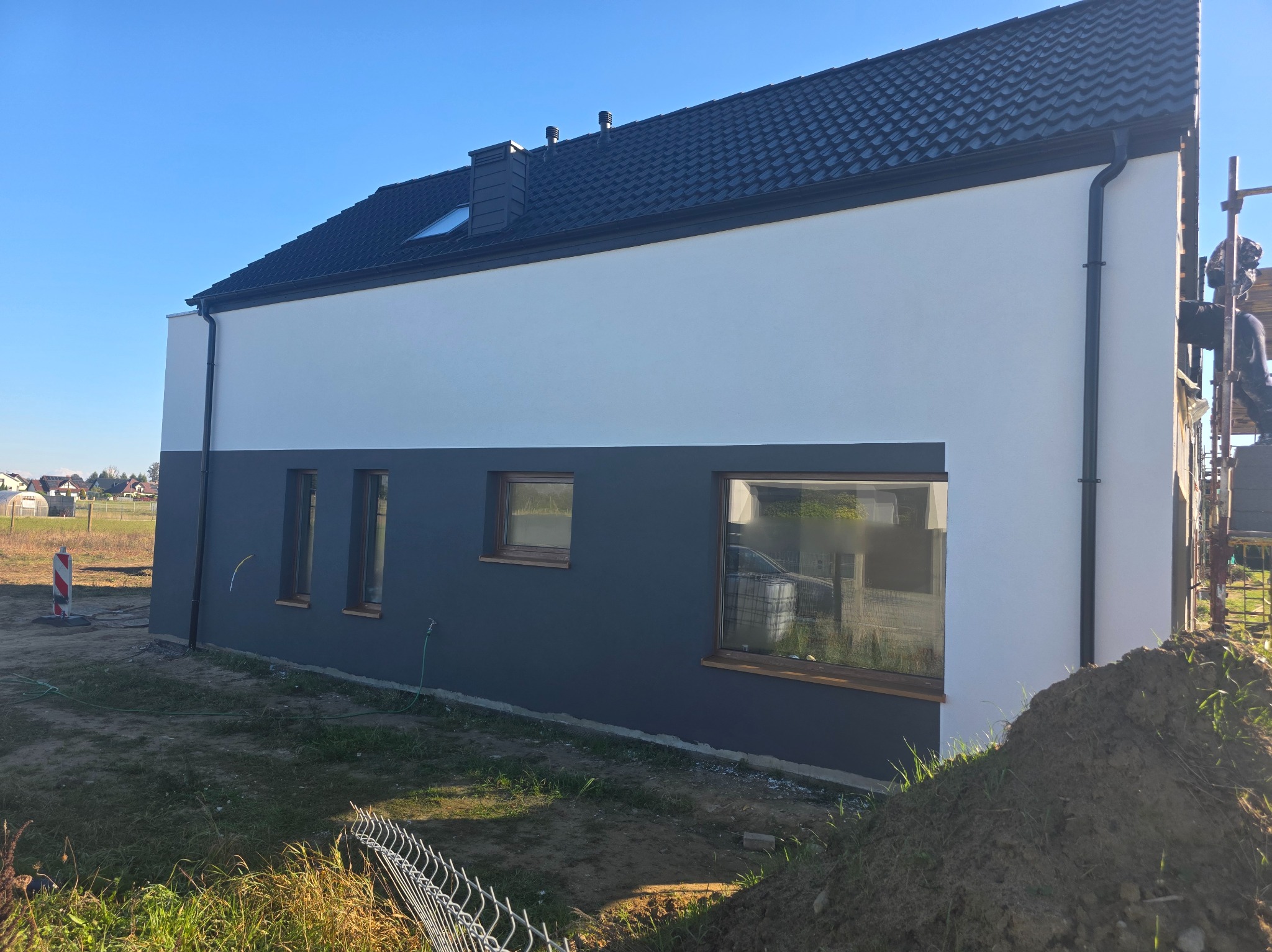 Dwukolorowa elewacja domu jednorodzinnego w odcieniach szarości i bieli, z widocznymi oknami i fragmentem rusztowania. Nowoczesny styl, słoneczny dzień.