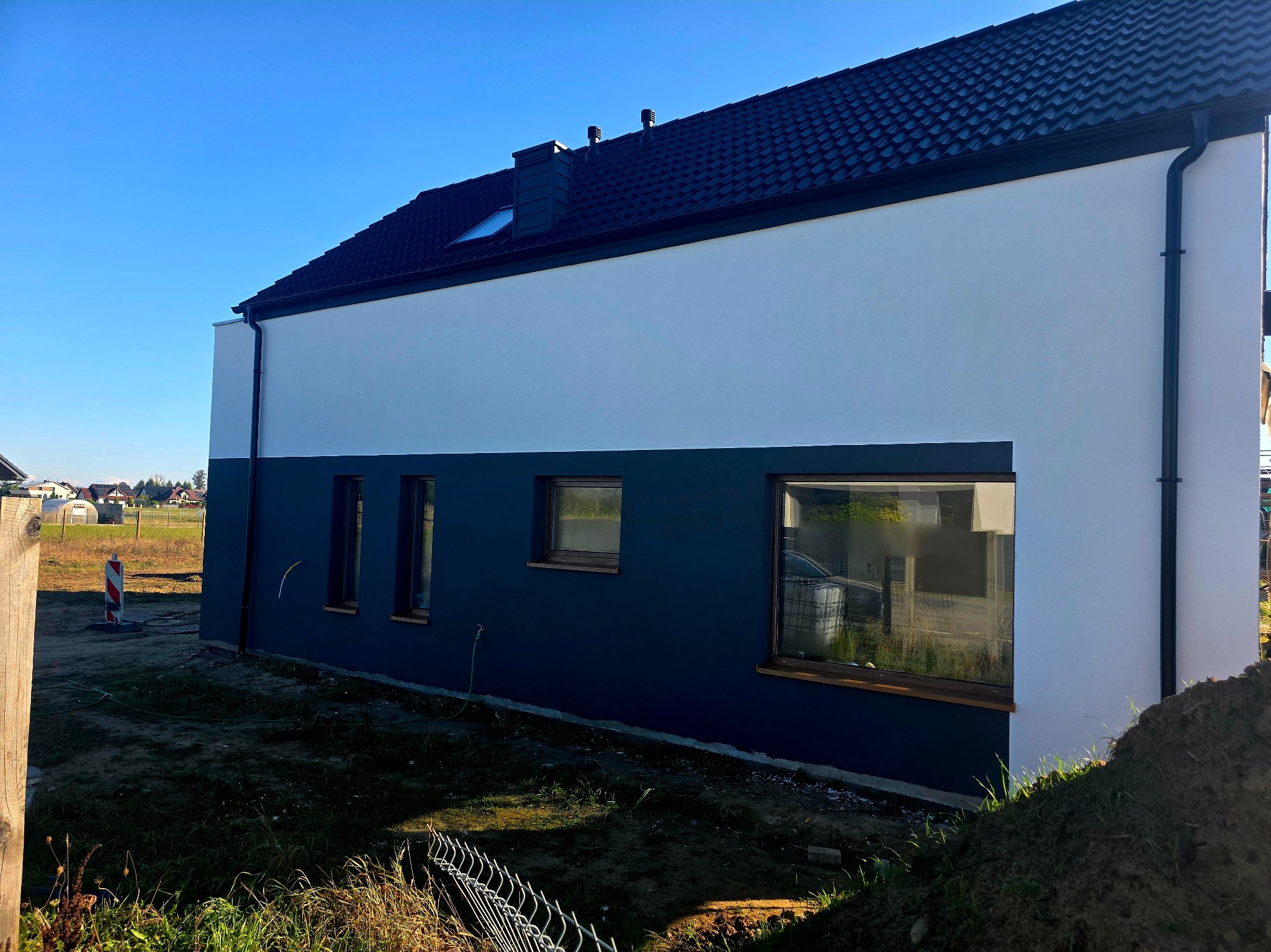 Dwukolorowa elewacja domu jednorodzinnego z czarną dachówką i drewnianą stolarką okienną. Widok z boku na tle błękitnego nieba i luźnej zabudowy.