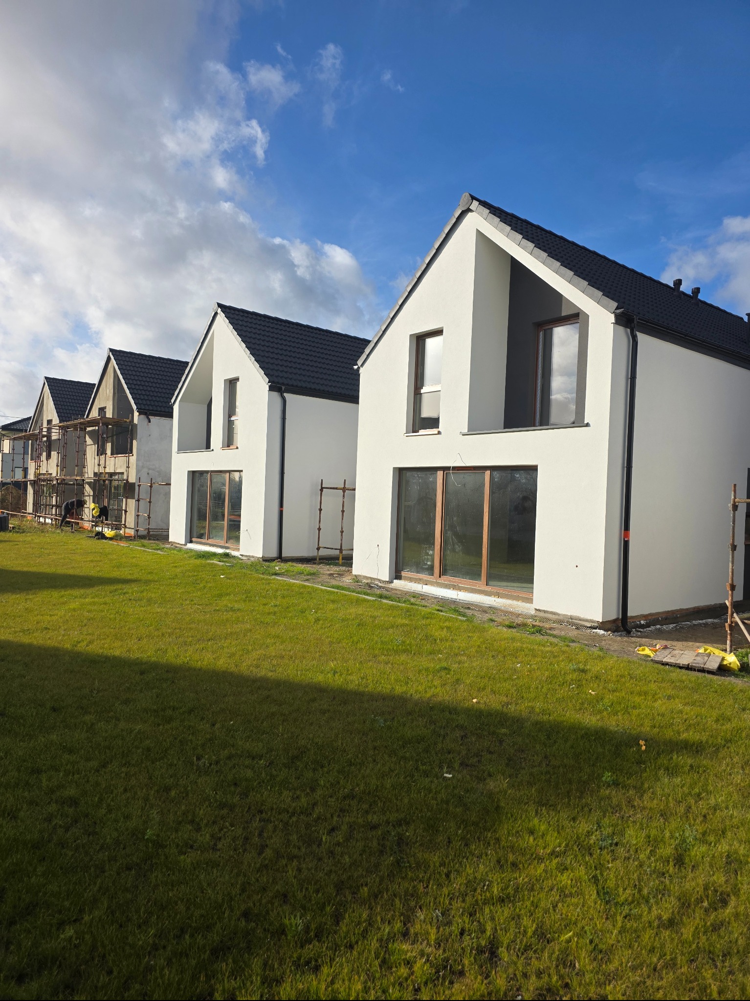 Nowoczesne domy jednorodzinne z białymi elewacjami i ciemnymi dachami, jeden dom w trakcie prac elewacyjnych, widoczne rusztowania i trawnik.