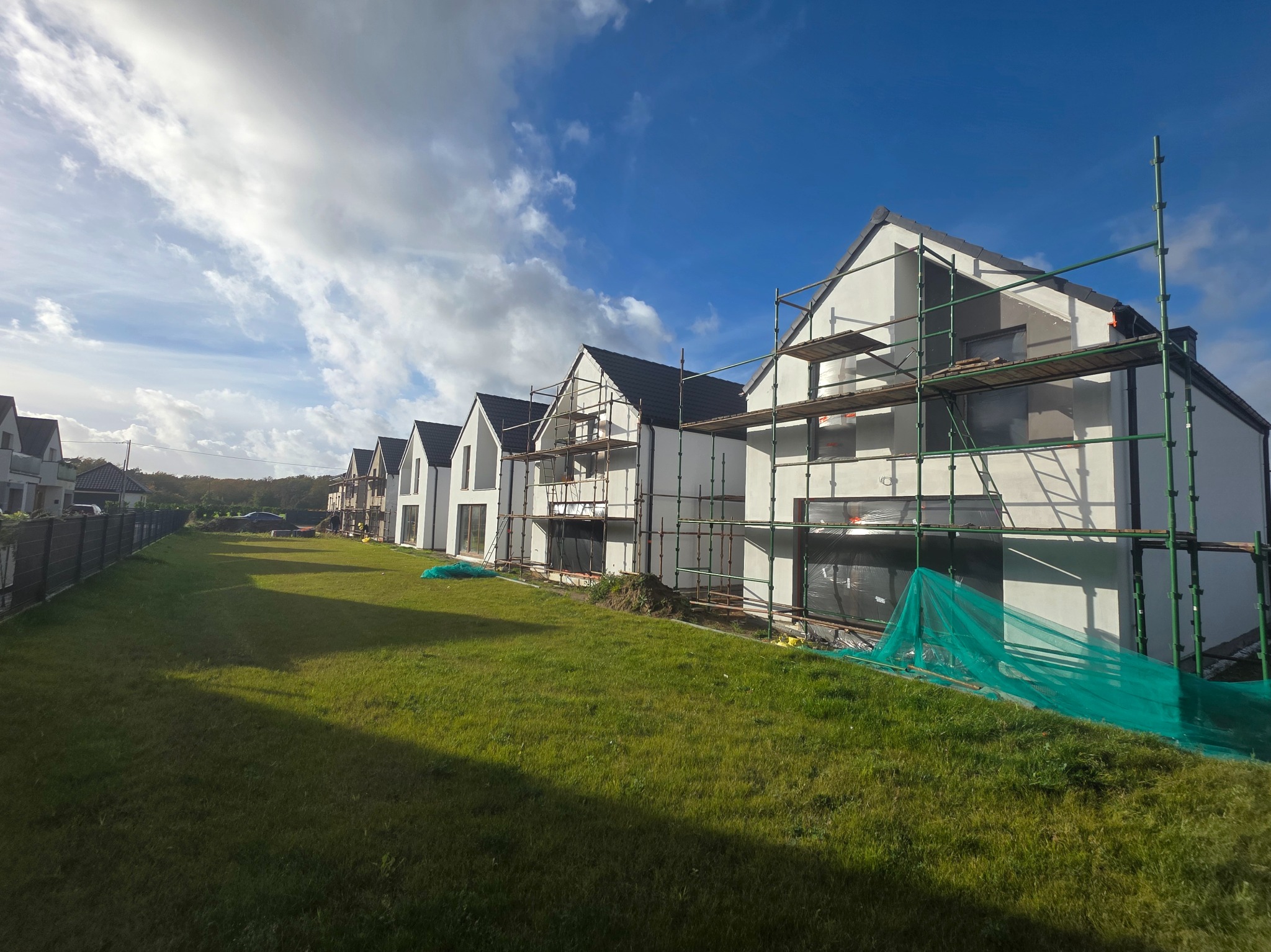 Rząd domów w trakcie prac elewacyjnych, widoczne rusztowania i zabezpieczenia, trawnik, błękitne niebo z chmurami w tle. Prace wykończeniowe elewacji.