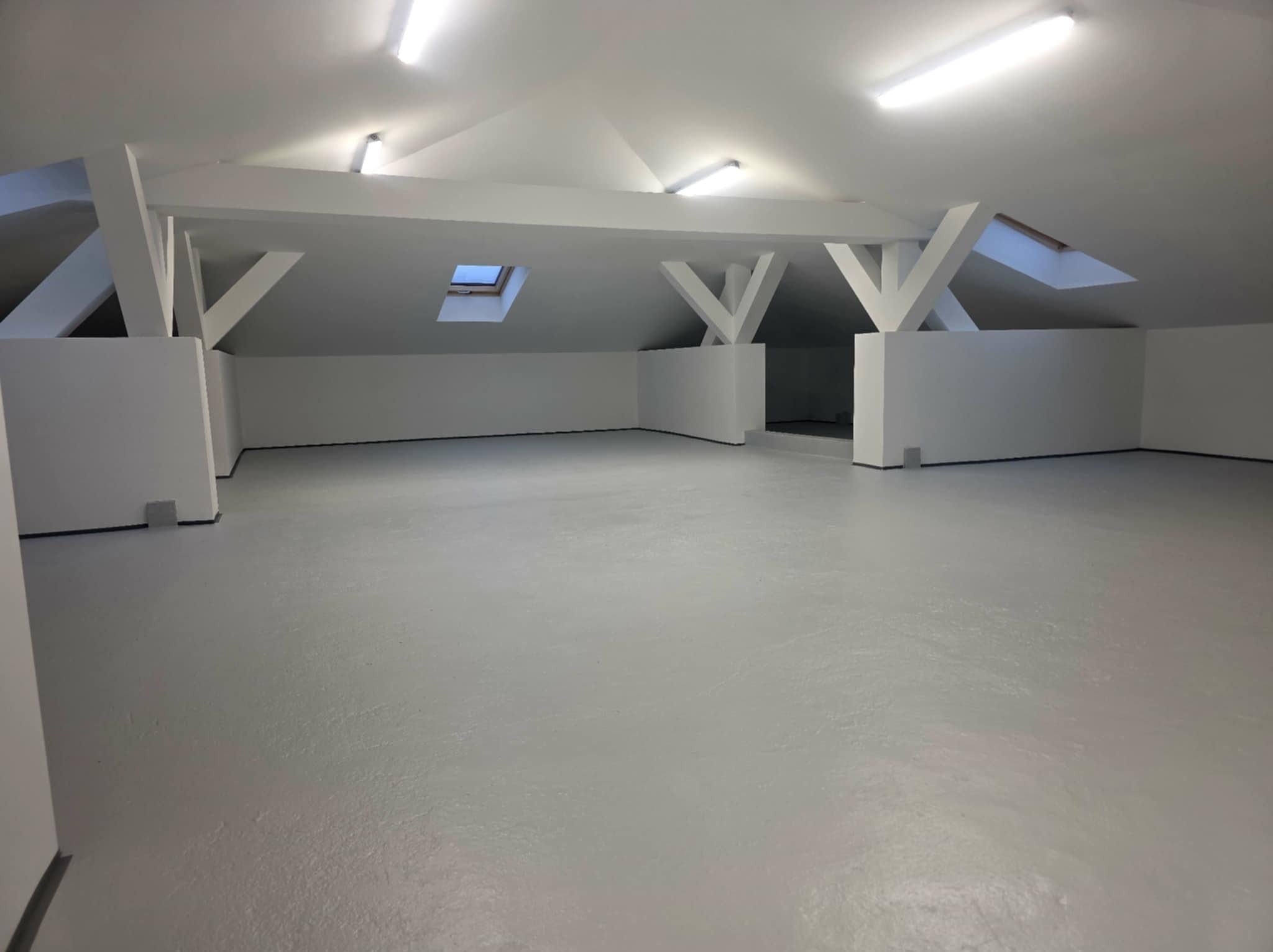 Pomalowane na biało poddasze ze skosami i oknami dachowymi w Połczynie. Widoczne drewniane elementy konstrukcyjne i gładka, jasna podłoga.