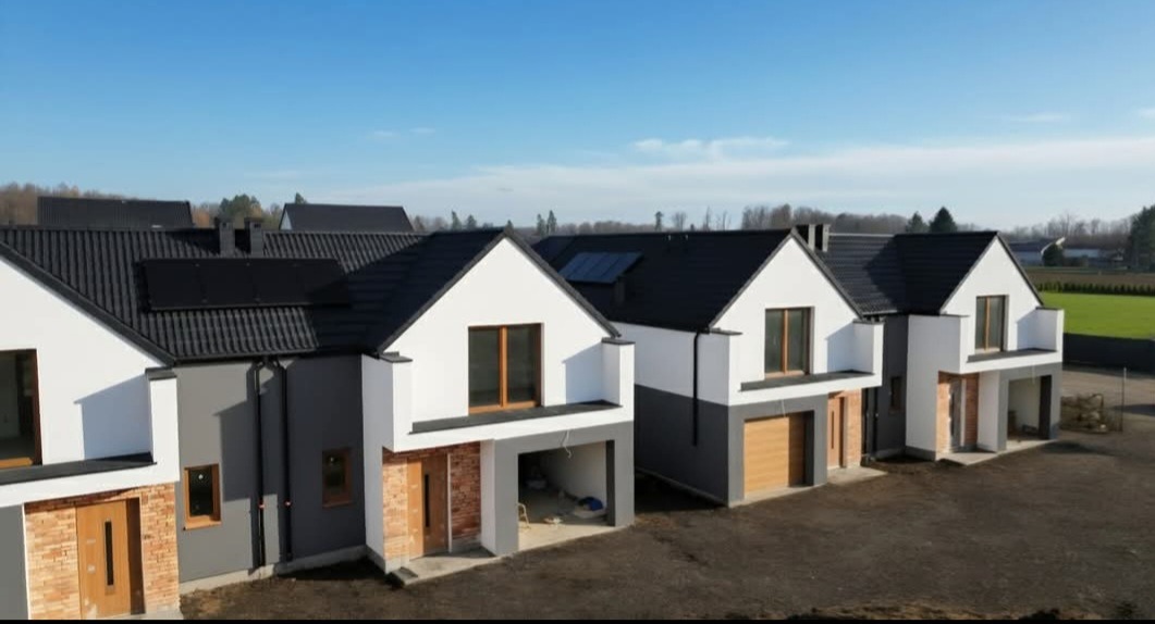 Rząd nowoczesnych domów jednorodzinnych z białymi elewacjami i ciemnymi dachami, niektóre z panelami słonecznymi, w słoneczny dzień. Widoczne garaże i elementy wykończeniowe.