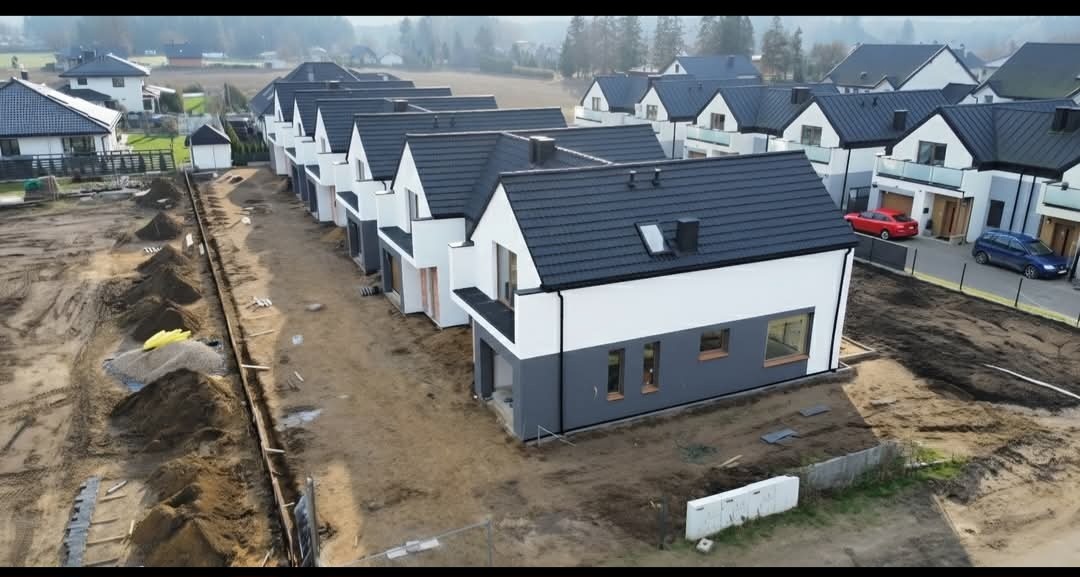 Nowe domy w zabudowie szeregowej z ciemnymi dachami i biało-szarą elewacją, widok z góry. Teren wokół domów w trakcie zagospodarowania, widoczne hałdy ziemi.