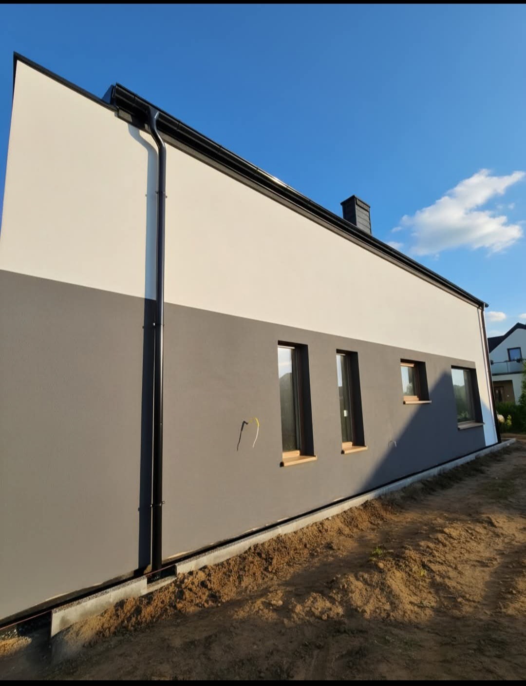 Dwukolorowa elewacja domu jednorodzinnego z czarną rynną i oknami, widoczna z poziomu gruntu w słoneczny dzień. Dolna część elewacji szara, górna biała.