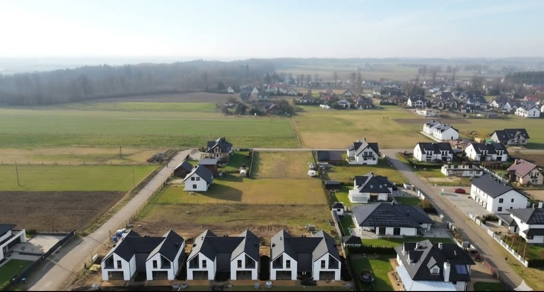 Osiedle nowych domów jednorodzinnych z czarnymi dachami, widok z lotu ptaka na tle pól i lasów. Domy w stylu nowoczesnej stodoły, w trakcie budowy.