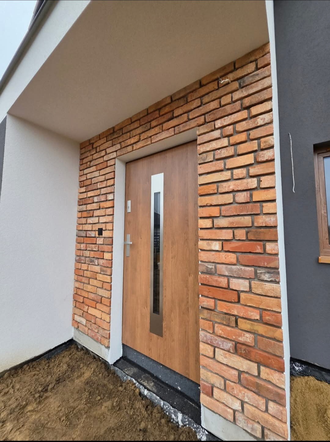 Elewacja domu z cegły klinkierowej wokół drewnianych drzwi z szybą. Szara ściana obok i biały fragment elewacji. Widoczny fragment gruntu przed wejściem.