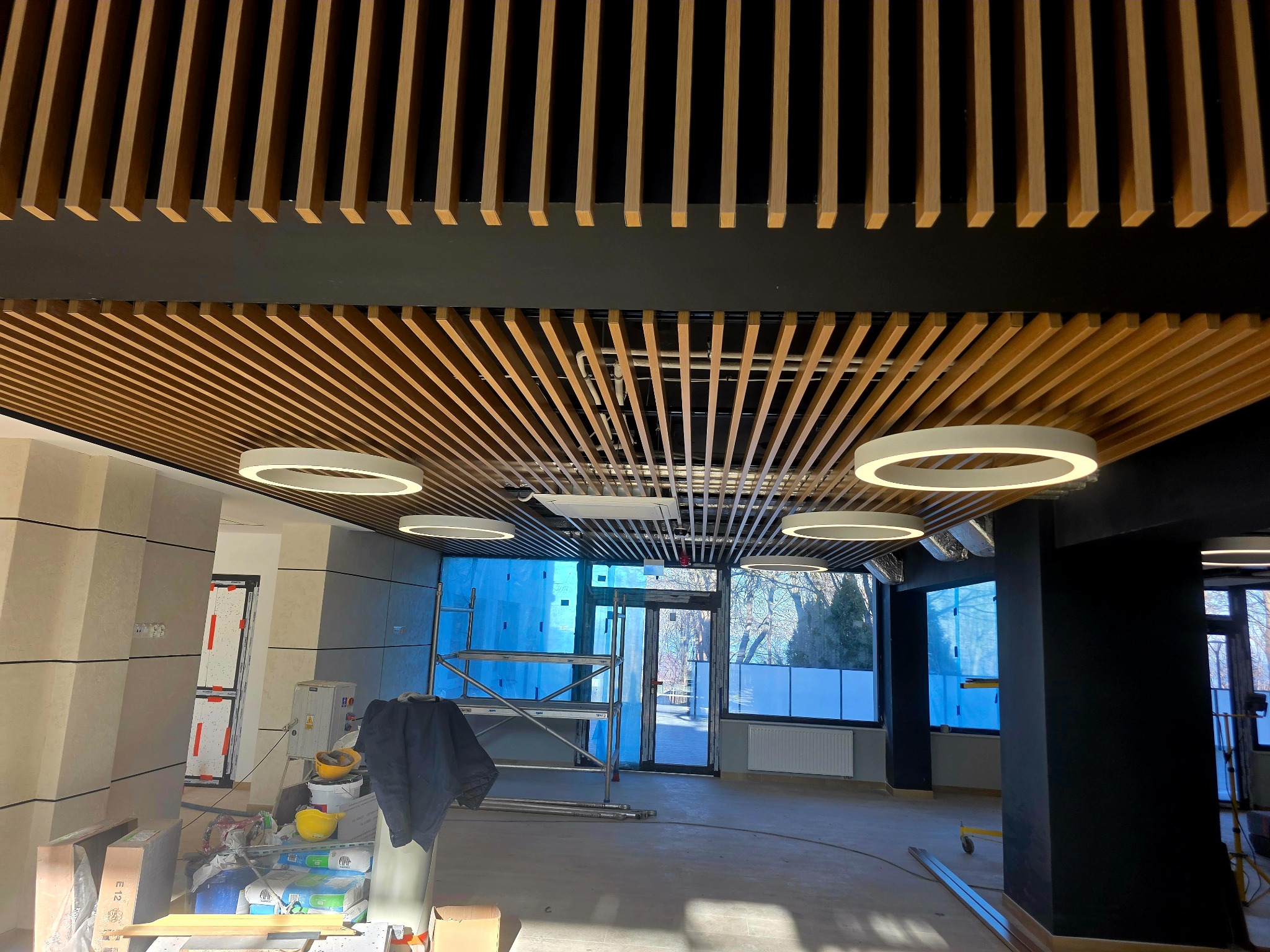 Nowoczesne wykończenie wnętrza biura z drewnianą ażurową konstrukcją sufitową i okrągłymi lampami. Widoczne rusztowania i materiały budowlane.