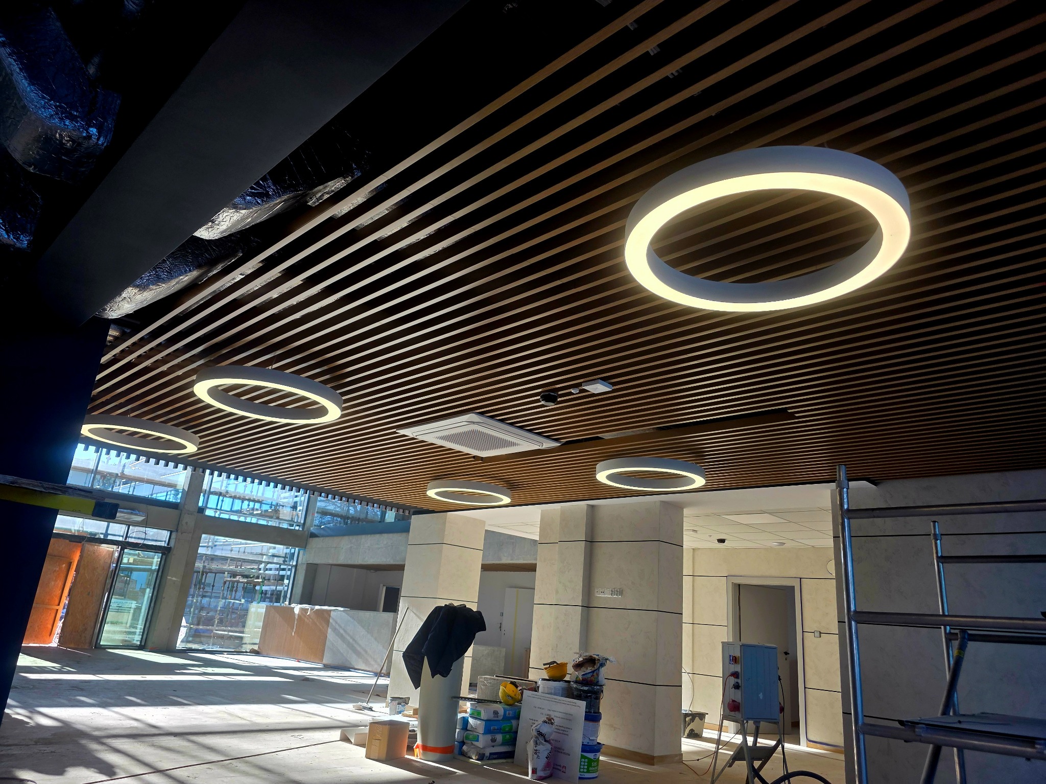 Montaż sufitu rastrowego z oświetleniem LED w kształcie pierścieni. Widoczne materiały budowlane i rusztowanie, co sugeruje etap wykończeniowy wnętrza komercyjnego.