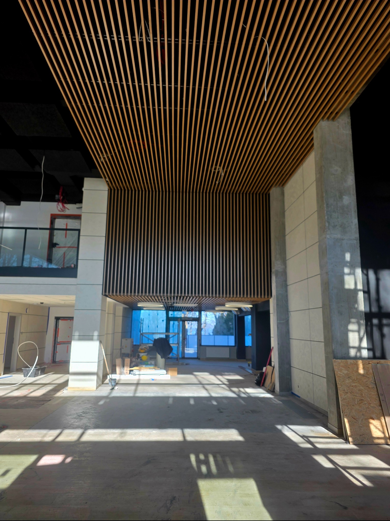 Wnętrze w trakcie wykończenia: drewniane lamele sufitowe i ścienne, surowy beton, jasne płytki. Widoczne elementy instalacji i narzędzia. Duże okna w tle.