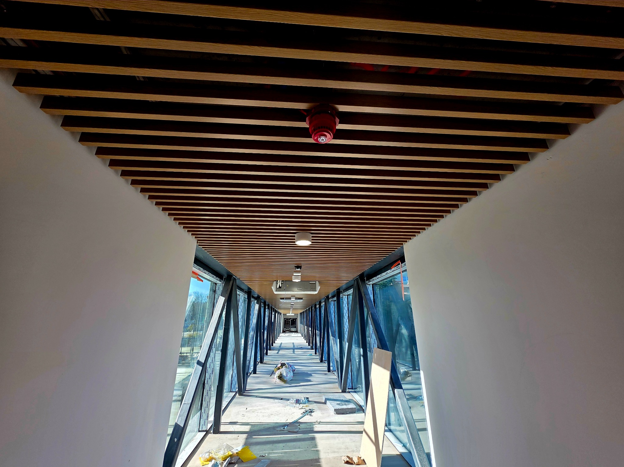 Wnętrze korytarza z drewnianym sufitem lamelowym i przeszklonymi ścianami, w trakcie prac wykończeniowych. Widoczne narzędzia i materiały budowlane.