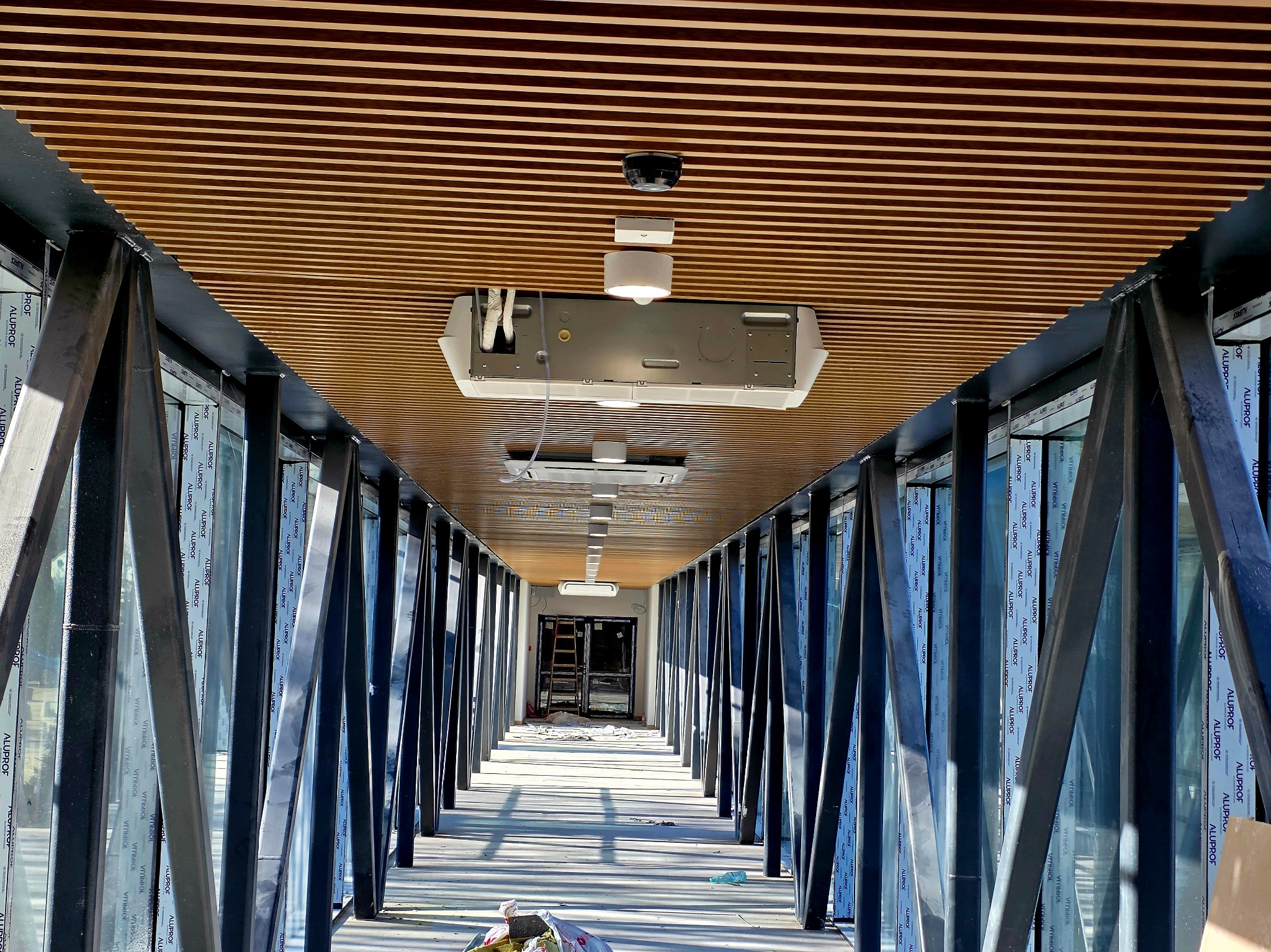 Wnętrze korytarza z drewnianym sufitem lamelowym, przeszklonymi ścianami z czarnymi słupkami i widoczną instalacją klimatyzacji pod sufitem.