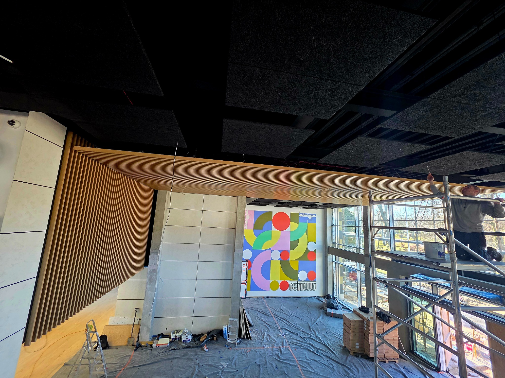 Wnętrze w trakcie wykończenia: drewniane lamele na ścianie, kolorowa grafika na ścianie, pracownik na rusztowaniu montuje panele akustyczne na suficie.