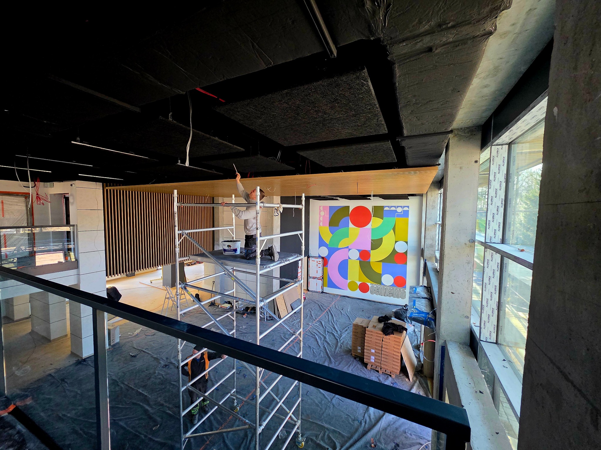 Prace wykończeniowe w nowoczesnym wnętrzu: malowanie sufitu z rusztowania, widoczna kolorowa grafika na ścianie i duże okna z widokiem na zewnątrz.