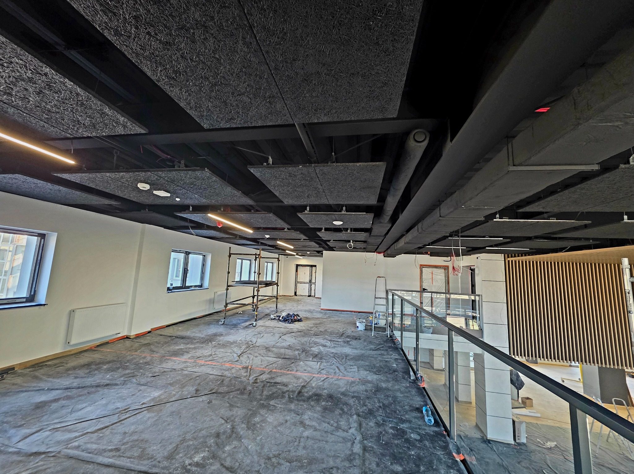 Wnętrze w trakcie wykończenia: zabezpieczona podłoga, rusztowania, widoczne elementy instalacji pod sufitem. Białe ściany, duże okna. Prace wykończeniowe w toku.