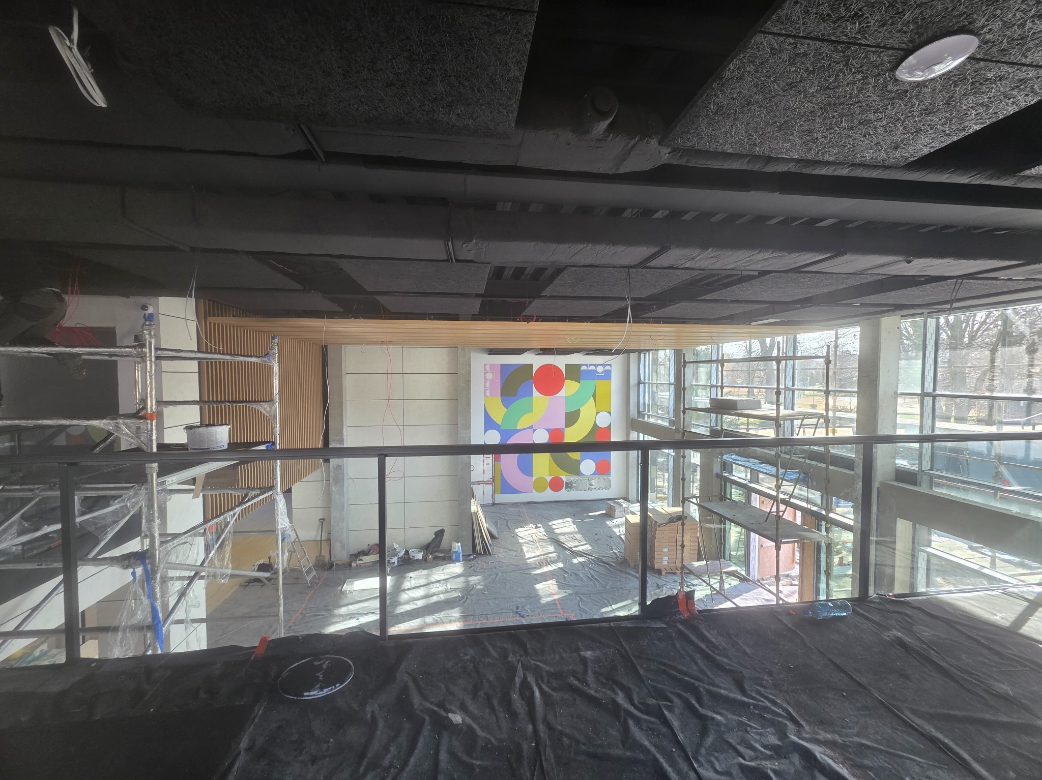 Wnętrze w trakcie wykończenia: sufit z panelami akustycznymi, ściana z kolorową grafiką, rusztowania i zabezpieczenia podłogi. Widok z góry.