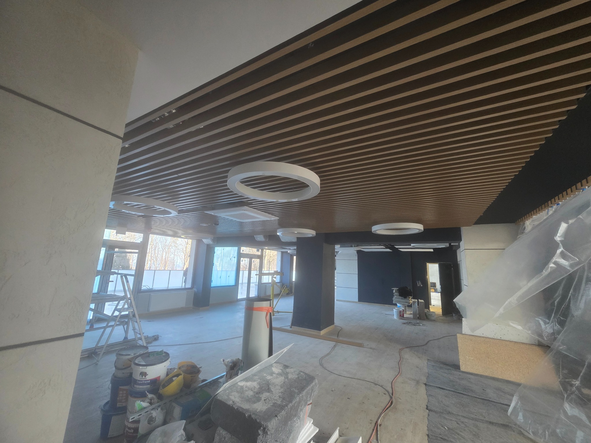 Wnętrze w trakcie wykończenia: drewniany sufit lamelowy z okrągłymi lampami, widoczne farby, drabina i zabezpieczenia. Duże okna wpuszczają naturalne światło.