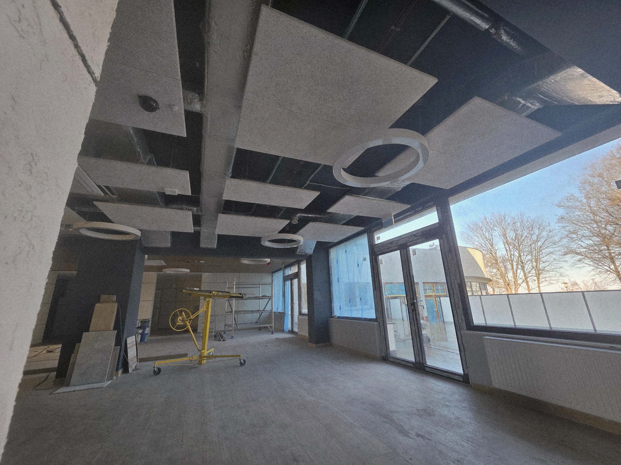 Wnętrze w trakcie wykończenia: sufit z panelami akustycznymi, duże okna z widokiem na zewnątrz, żółty podnośnik do płyt. Widoczne materiały budowlane i rusztowania.