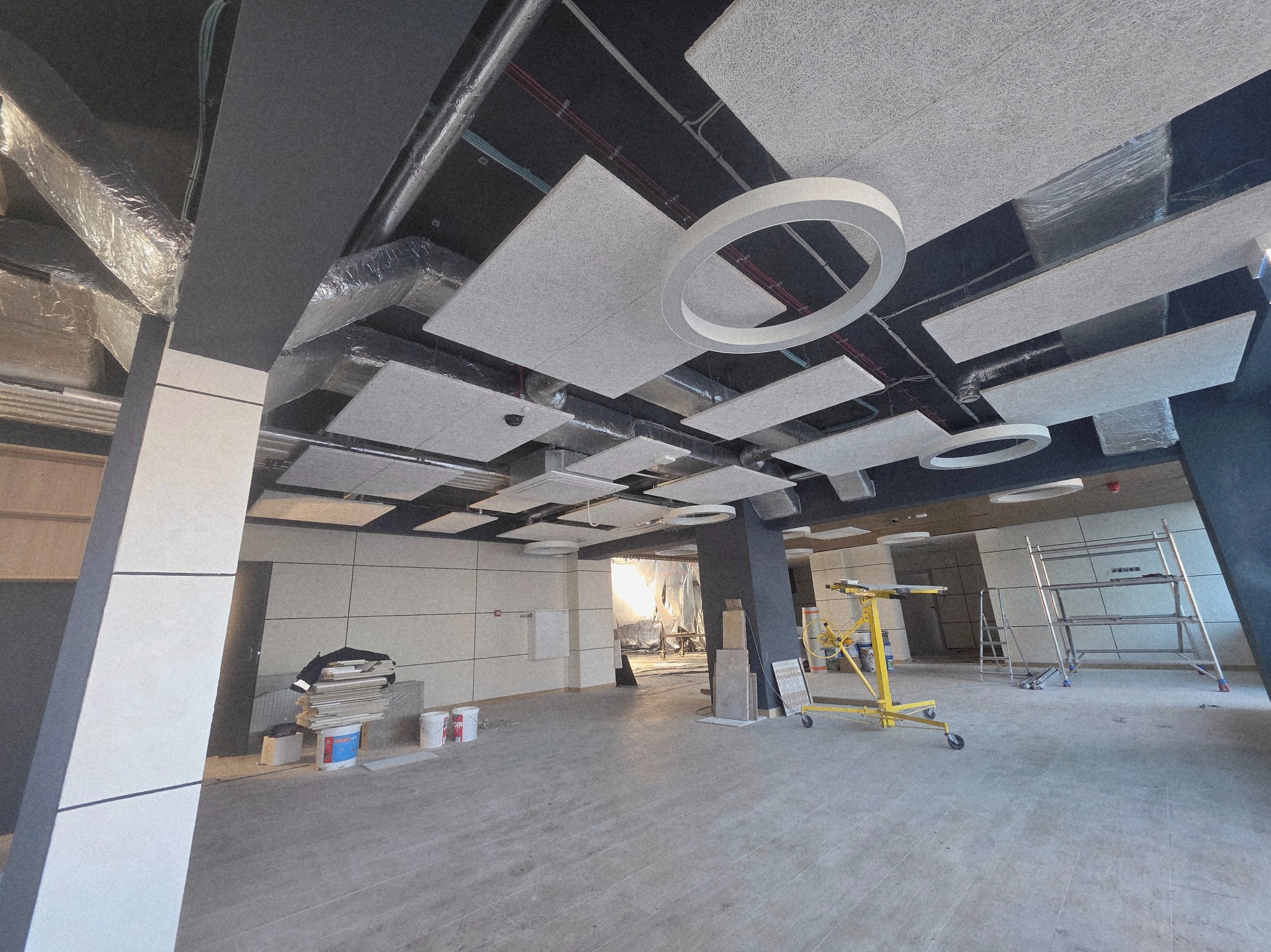 Montaż sufitu podwieszanego z paneli akustycznych w nowoczesnym biurze. Widoczne narzędzia i rusztowania. Jasne, przestronne wnętrze w trakcie wykończenia.