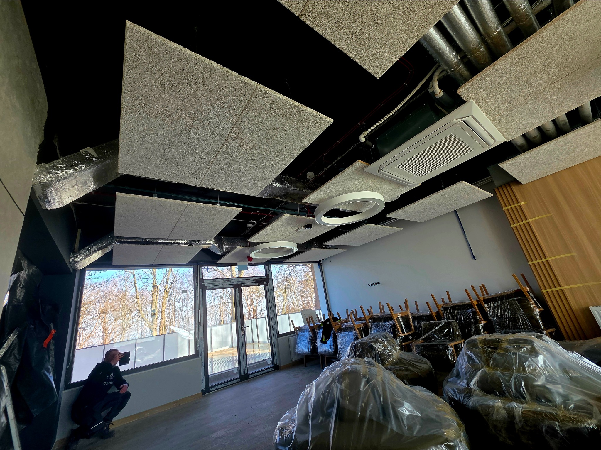 Montaż paneli akustycznych na suficie w nowym biurze. Widoczne instalacje, oświetlenie i pracownik przy oknie. Krzesła w folii czekają na ustawienie.