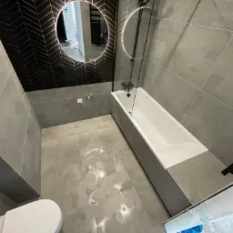 Nowoczesna łazienka z szarą glazurą, czarną ścianą z wzorem chevron, okrągłym lustrem z podświetleniem LED, białą wanną z parawanem i wiszącą toaletą.