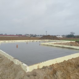 Usługi Budowlane DEKORBUD - Zalany fundament z izolacją w Piotrków Kujawski. Widoczne wystające rury i piasek. W tle pole i zabudowania.