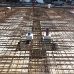 Siatka zbrojeniowa ułożona na przygotowanym podłożu, widoczne pionowe rury z niebieskimi zaślepkami, przygotowane do wylania betonu.