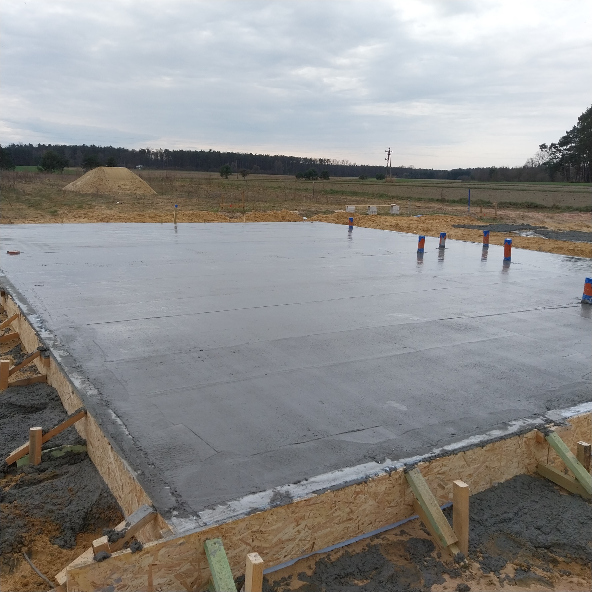 Wylana płyta fundamentowa z widocznym szalunkiem i wystającymi rurami instalacyjnymi na tle krajobrazu z lasem i stertą piasku. Widok z poziomu gruntu.