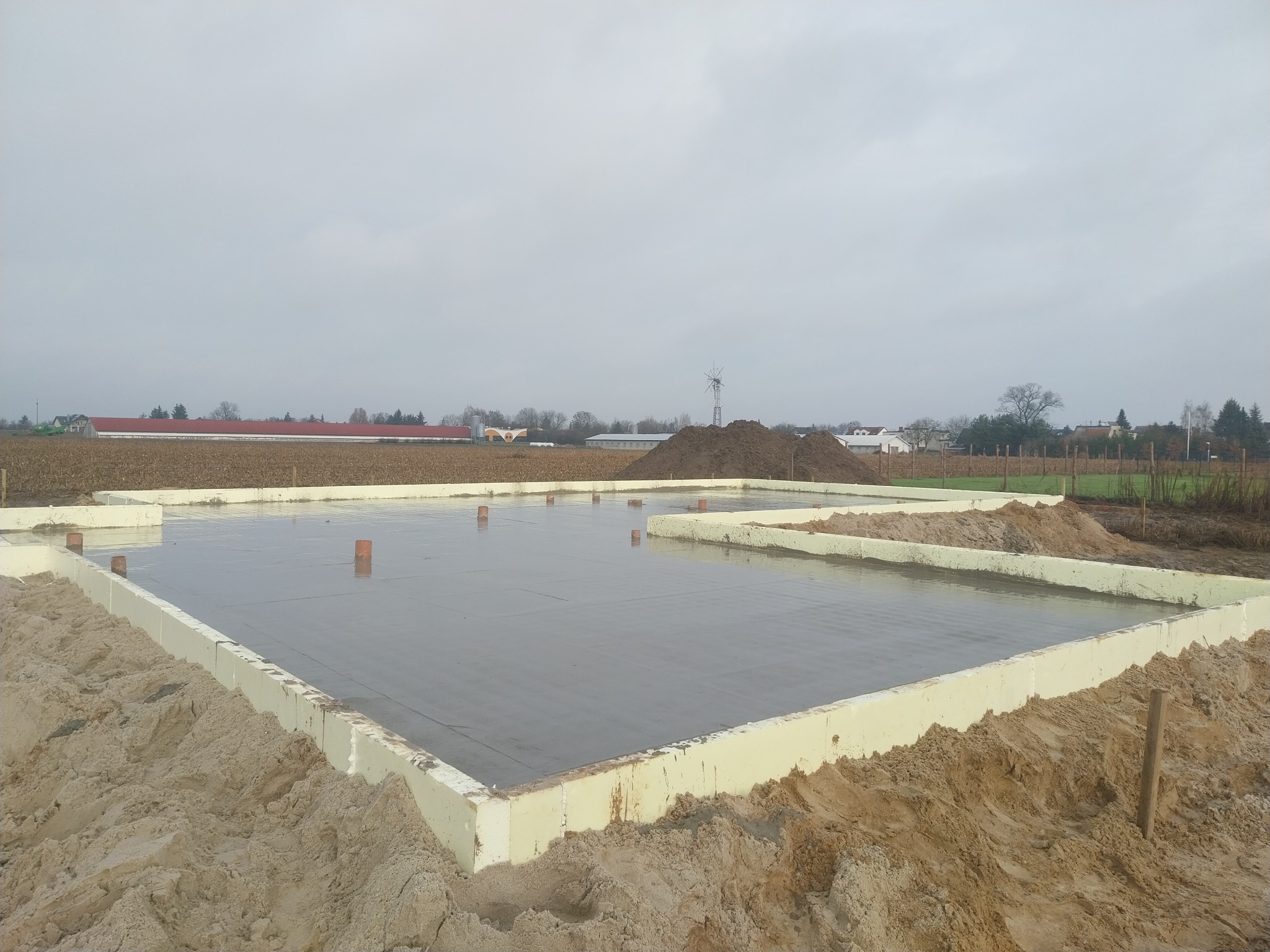 Zalany fundament z izolacją w Piotrków Kujawski. Widoczne wystające rury i piasek. W tle pole i zabudowania.
