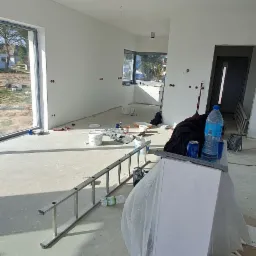 Wnętrze w trakcie prac wykończeniowych: świeżo pomalowane, białe ściany, widoczne puszki, drabina, wiadro i inne akcesoria malarskie na podłodze, niedokończone gniazdka elektryczne.