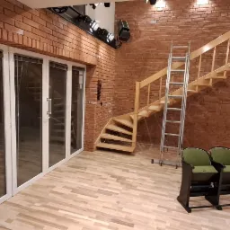 Wnętrze z ceglaną ścianą, drewnianymi schodami, metalową drabiną i rzędem kinowych foteli na parkiecie.