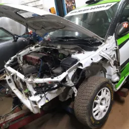 Uszkodzony samochód rajdowy z otwartą maską, widoczne elementy silnika i opony z bieżnikiem rajdowym, karoseria w trakcie naprawy na podnośniku.