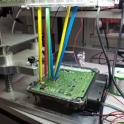 Elektroniczna jednostka sterująca silnika (ECU) podłączona do przewodów diagnostycznych w kolorach żółtym, niebieskim, zielonym i czerwonym, na stole warsztatowym z widocznym oprzyrządowaniem...