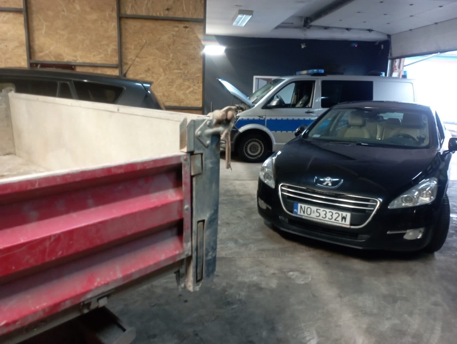 Część skrzyni ładunkowej samochodu ciężarowego na tle policyjnego samochodu i czarnego osobowego Peugeota z otwartą maską w warsztacie.