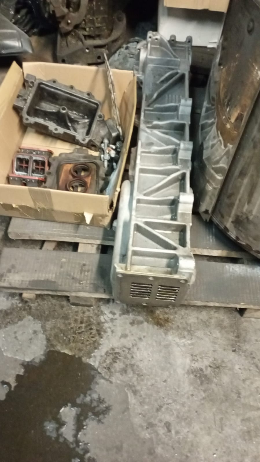 Wyjęte elementy silnika samochodowego, w tym aluminiowa obudowa skrzyni biegów i części zdemontowane do naprawy, umieszczone w kartonowym pudle na palecie w warsztacie.