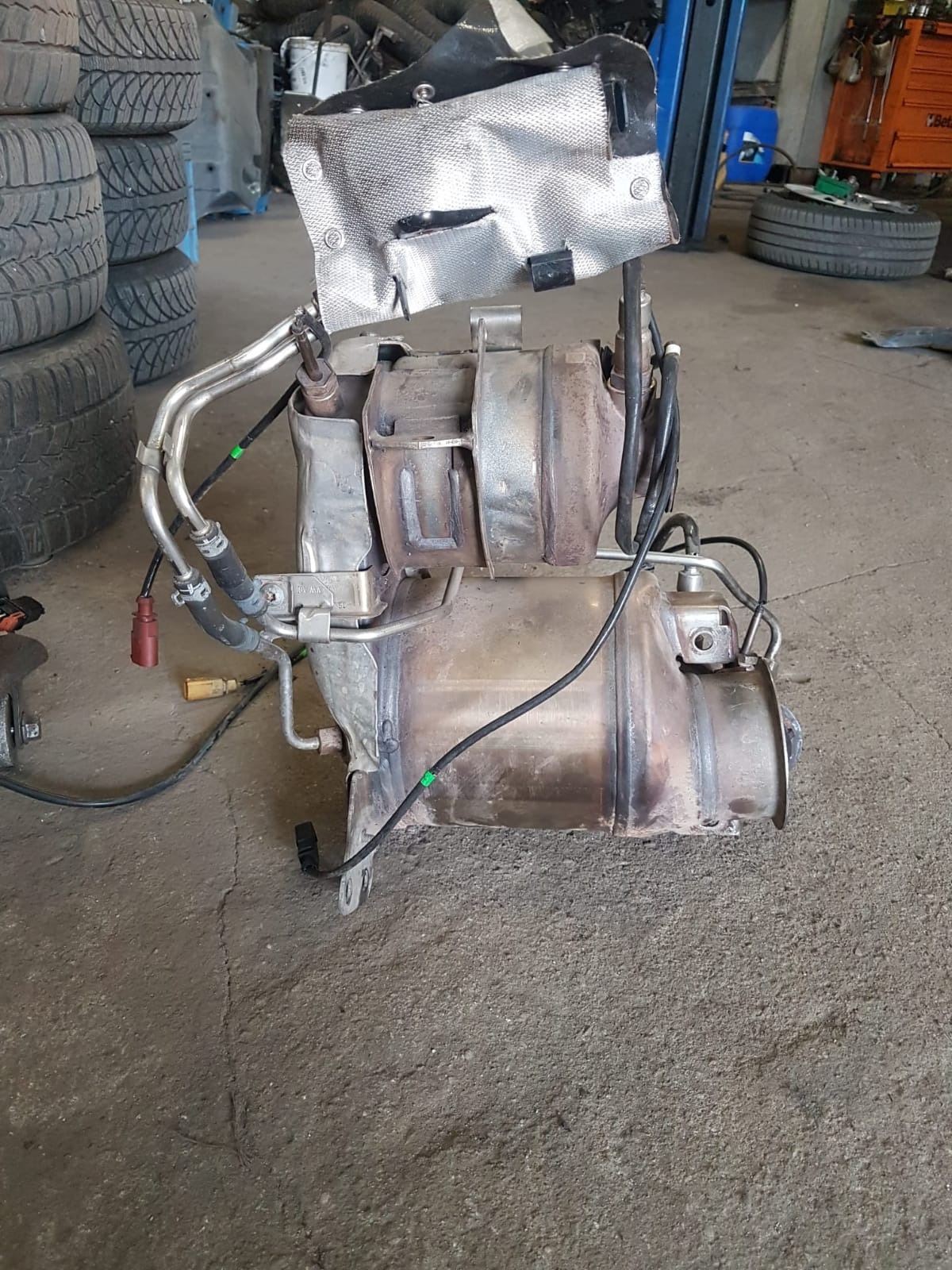 Wymontowany, używany filtr cząstek stałych (DPF) z osłoną termiczną, widoczne przewody i czujniki, na betonowej posadzce warsztatu, w tle opony i narzędzia.