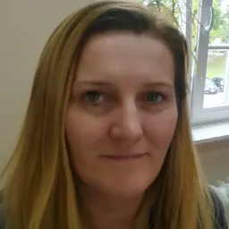 Portret kobiety o blond włosach na tle okna z widokiem na zieleń i zaparkowane samochody.