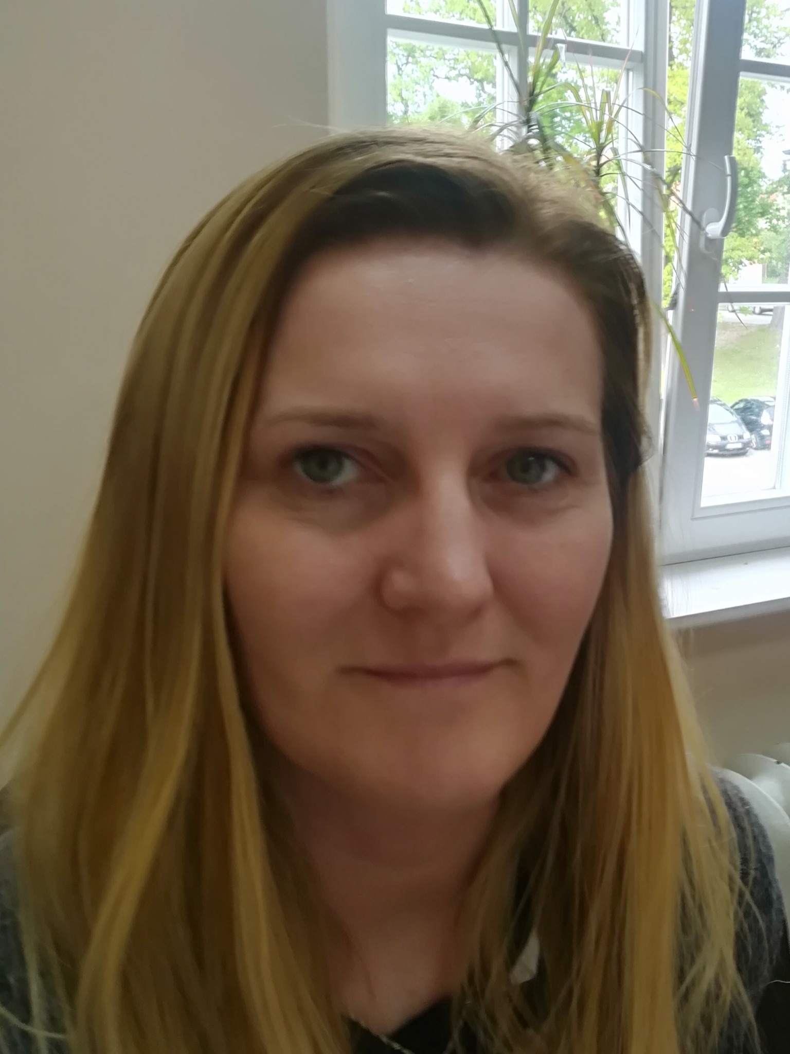 Portret kobiety o blond włosach na tle okna z widokiem na zieleń i zaparkowane samochody.